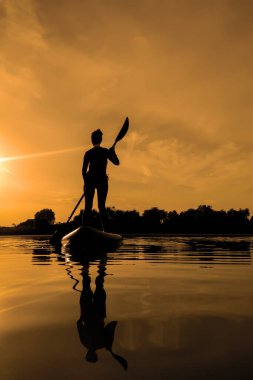 Yemek masasında bir kadın, gün batımında sörf yaparken suda yansıyor.