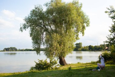 Beyaz elbiseli ve hasır şapkalı genç bir kadın gölün kenarındaki ahşap bankta oturuyor.