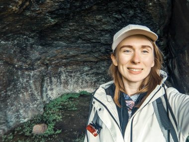 Sırt çantalı gülümseyen kadın kaya arka planına selfie çekiyor.