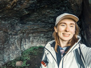 Sırt çantalı gülümseyen kadın kaya arka planına selfie çekiyor.