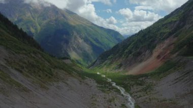 Aerial view of a mountain stream flowing through a valley between cloud-covered mountain peaks against a cloudy blue sky on a summer day. Clouds cast shadows on the mountains.
