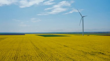 Güzel bir bahar gününde, sarı kolza tarlalarında uçan bir yel değirmeninin hava görüntüsü. Çevre koruma. Yenilenebilir alternatif enerji kaynakları.