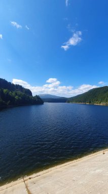 Barajdan görünen Oasa Gölü. Uzaktaki yeşil orman dağları. Mavi gökyüzüne dağılmış bulutlar. Güzel bir yaz günü. Oasa, Romanya