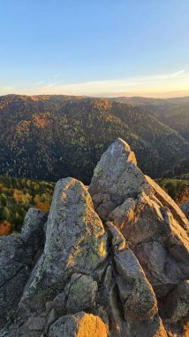 Gün batımında renkli ormanlarla dağların tepesinden vadiye doğru kayaların manzarası. Ağaçlarda sarı, turuncu ve yeşil karışımlar, açık mavi gökyüzü. Marisel, Cluj, Romanya