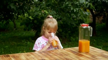 Little girl drinks orange juice in the garden. A child drinks orange juice while sitting at a table against the backdrop of plants. Slow motion.