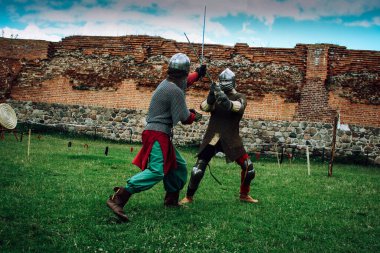 Miğferli iki şövalyenin kılıç dövüşü ve ortaçağ kalesi manzaralı zırhlı zırhlar.