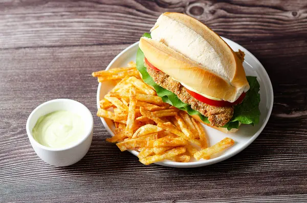 Domates dilimli Milanesa sandviçi, marul ve mayonez ve beyaz tabakta patates kızartması, ve ahşap zemin üzerinde mayonez olan bir kase..