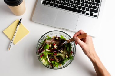 Salatada çatal, dizüstü bilgisayar, kalem ve yapışkan notlar ve yeniden kullanılabilir kahve tutan bir kadın eli olan bir işyeri manzarası. İş yerinde sağlıklı yiyecek kavramı. 