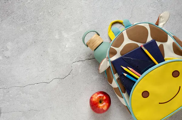 Okul malzemeleriyle dolu zürafa çantası, beton yüzeyde kırmızı elma ve su şişesi. Okul konseptine dönelim.. 