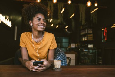 Kulaklıklı gülümseyen genç bir kadın rahat bir kafede oturup kahvenin tadını çıkarırken akıllı telefon kullanıyor.