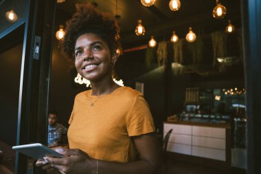 Gülümseyen genç bayan kafe sahibi elinde dijital tabletle modern kahve dükkanının girişine bakıyor.