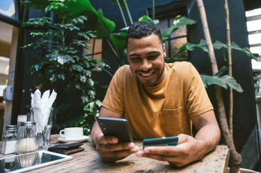 Akıllı telefon ve kredi kartı kullanarak çevrimiçi ödeme yapan gülümseyen genç adam rahat bir kafedeki ahşap masada otururken...