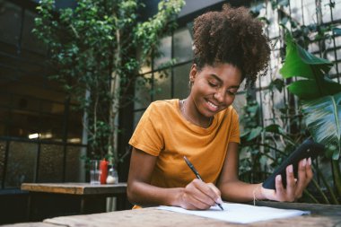 Akıllı telefon kullanan ve kapalı kafedeki bitkilerle dolu ahşap masaya notlar yazan gülümseyen genç bir kadın.