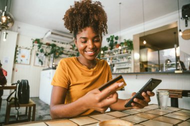 Akıllı telefon ve kredi kartı kullanarak çevrimiçi ödeme yapan gülümseyen genç bir kadın rahat bir kafedeki ahşap masada otururken...