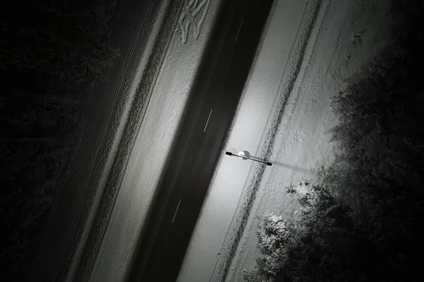 An atmospheric drone photo of a winter road winding through a snow-covered forest in mountains at night. The trees are adorned with a delicate layer of hoarfrost, creating a magical winter wonderland