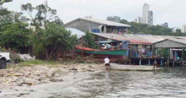 Sihanoukville - Kamboçya - 07- 12- 2023; iki adam küçük bir tekneyi enkazla dolu küçük bir sahile ittiler. Arka planda balıkçı köyü