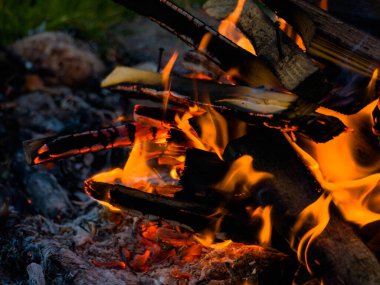 Küçük bir yığın halinde kesilmiş odun parçaları. Üzerlerinde yanan şenlik ateşinin alevleri uçuşuyor. Ahşap siyahtır ve bir kül tabakasıyla kaplıdır..