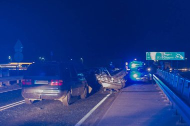 Geceleri asfalt yol, araba farlarının ışığıyla aydınlatılıyor. Yolda bir trafik kazasına karışan iki kırık araba görebilirsiniz, acil servislerin yanıp sönen mavi ışıklarını görebilirsiniz..