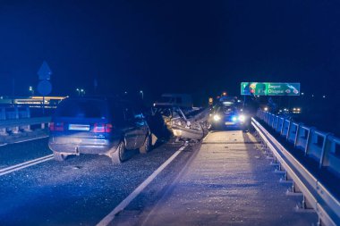 Geceleri asfalt yol, araba farlarının ışığıyla aydınlatılıyor. Yolda bir trafik kazasına karışan iki kırık araba görebilirsiniz, acil servislerin yanıp sönen mavi ışıklarını görebilirsiniz..