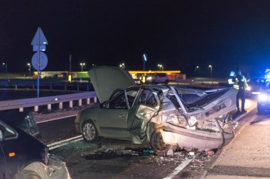 Geceleri asfalt yol, araba farlarının ışığıyla aydınlatılıyor. Yolda bir trafik kazasına karışan iki kırık araba görebilirsiniz, acil servislerin yanıp sönen mavi ışıklarını görebilirsiniz..