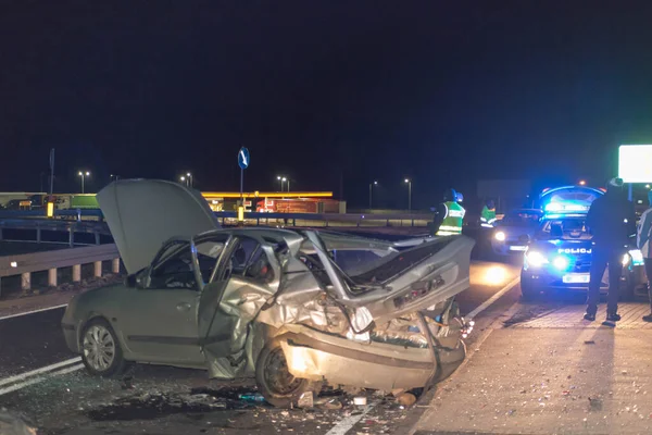 Geceleri asfalt yol, araba farlarının ışığıyla aydınlatılıyor. Yolda bir trafik kazasına karışan iki kırık araba görebilirsiniz, acil servislerin yanıp sönen mavi ışıklarını görebilirsiniz..