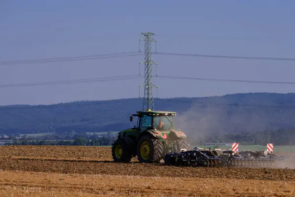 Sudeten foothills, undulating, uneven terrain covered with forests, meadows and arable fields. A tractor is working in a farm field with dust rising behind it.