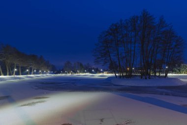 Gece vakti, kış manzarasında bir gölet. Göletin yüzeyi buzla kaplıdır, kıyı beyaz kar tabakasıyla kaplıdır. Gölün etrafında bir patika var. Banklar ve fenerler var. Bir İHA 'dan fotoğraf.