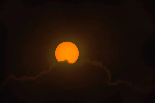 The solar disk photographed with a telephoto lens. As a result of using an optical filter, a warm color of the sun disc was obtained. Sun spots are visible on the surface. From below, the sun is partially obscured by clouds.
