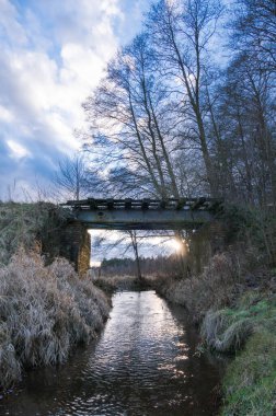 An old ruined railway bridge over a stream on a snowless winter afternoon. Sunlight breaks through the partly cloudy sky. clipart