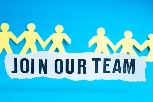 Join our team, written with stamped capital letters on a strip of white paper, with defocused yellow silhouettes on turquoise background
