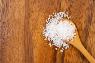 Wooden tea spoon with granulated salt spilling on textured wood background, flat lay close up