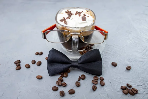 Glass cup of coffee with whipped cream and coffee beans. Coffee, bowtie and eyeglasses on cement gray texture.