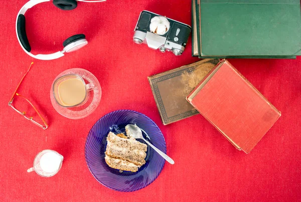 Vintage books, cake slice, coffee cup, headphones and glasses flat lay on red table cloth background
