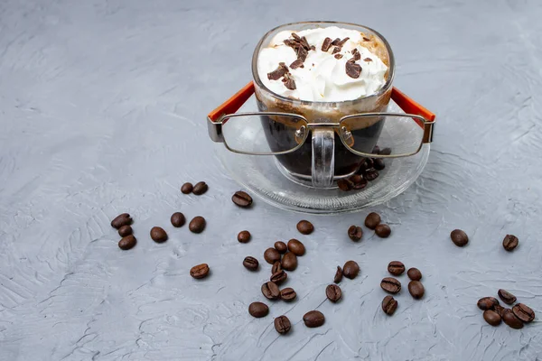 Glass cup of coffee with whipped cream and coffee beans. Coffee,   and reading eyeglasses on cement gray texture.