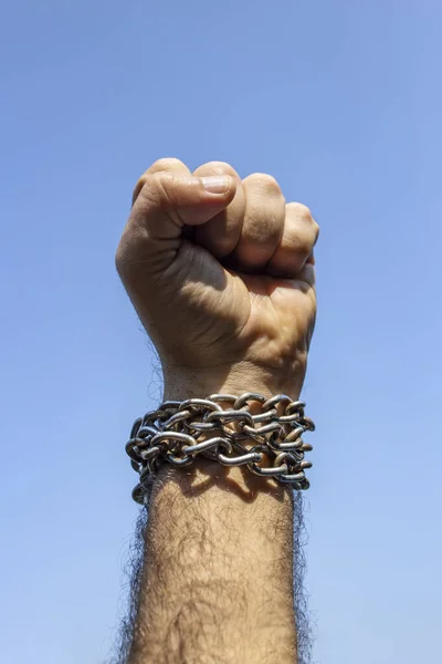 Clenched male fist raised with steel chain tied on the wrist, against blue sky
