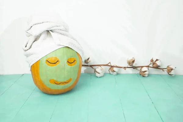 Orange pumpkin with a green facial mask, and a white towel on turquoise wooden boards table, empty space backdrop