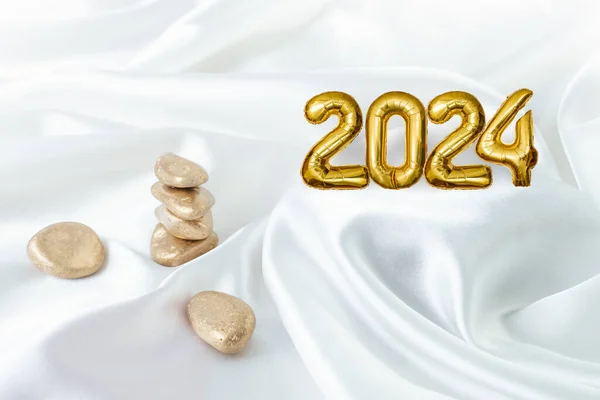 2024 tin foil balloons number and golden stones stack on white satin background. Zen garden concept.