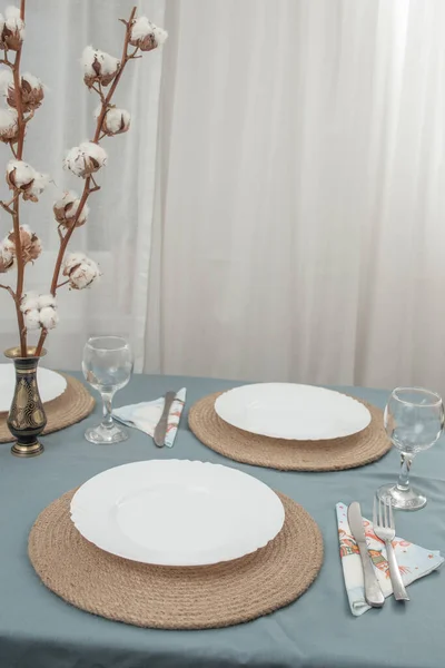 Table setting with jute rope placemats, white plates, cutlery, cotton flowers vase and blue tablecloth, side view