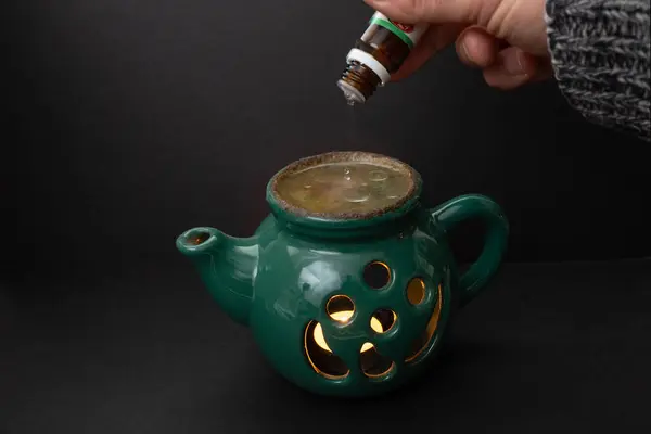 Candle essential oil burner in shape of a green tea pot, with a hand pouring a small bottle on dark background,