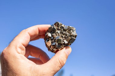 Erkek el mavi gökyüzüne karşı altın mineralli bir Pyrite taşı tutuyor, güneş ışığında altını kandırıyor.  