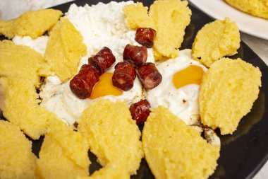 Polenta, sahanda yumurta, sosis ve siyah tabakta süzme peynir. 