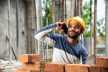 Mutlu gülümseyen genç Hintli inşaat işçisi iş yerinde cep telefonuyla meşgul.