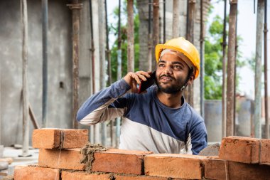 Mutlu gülümseyen genç Hintli inşaat işçisi iş yerinde cep telefonuyla meşgul.