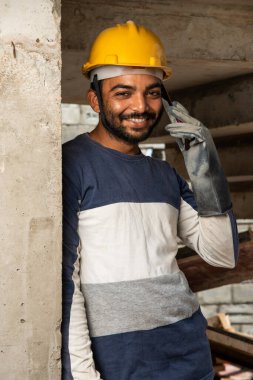 Mutlu gülümseyen genç Hintli inşaat işçisi iş yerinde cep telefonuyla meşgul.