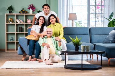 Modern evdeki oturma odasında grup fotoğrafı için poz veren dört kişilik Hintli aile.