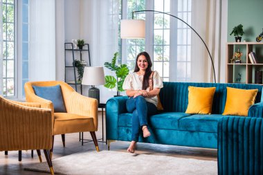 Modern oturma odasındaki kanepede oturan güzel Hintli genç kadın kameraya bakıyor.