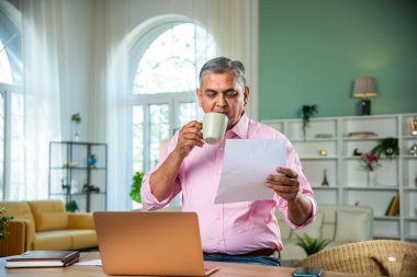 Hintli orta yaşlı adam dizüstü bilgisayarda çalışıyor, akıllı telefonlu ve belgeli kahve içiyor.