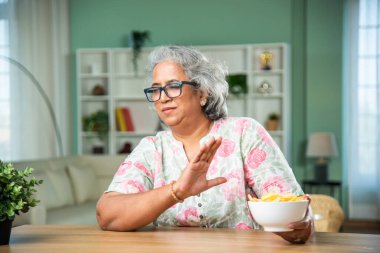 Unhealthy diet - senior indian woman refusing to eat potato chips from bowl clipart