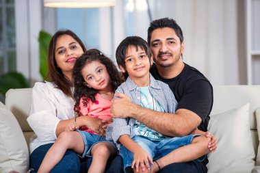 Oturma odasındaki kanepede oturan dört kişilik mutlu Hintli genç aile portresi.