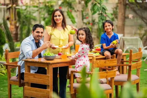 indian young family of four eating meal on dining table in the garden